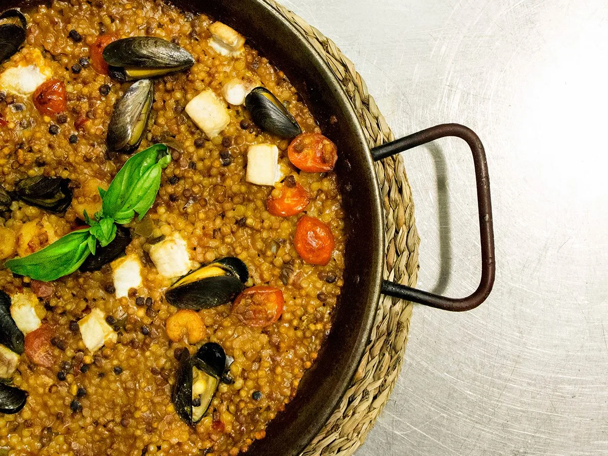 Fregola sarda con cozze, bocconcini di pesce, pomodorini e foglie di basilico in una padella di ferro su un sottopentola di paglia