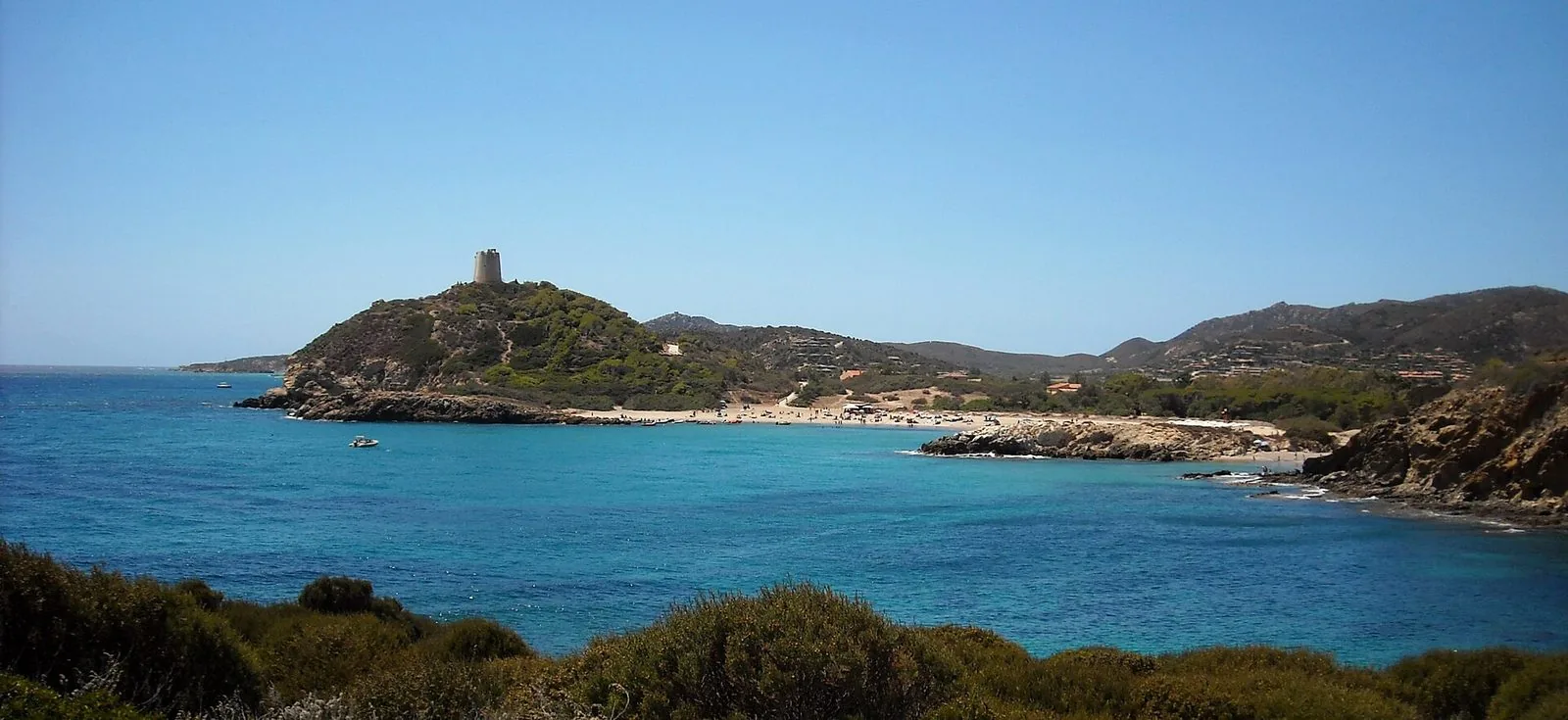 La costa di Chia con la torre di avvistamento sul promontorio verde, una piccola spiaggia di sabbia chiara, il mare turchese e in primo piano i cespugli della macchia mediterranea