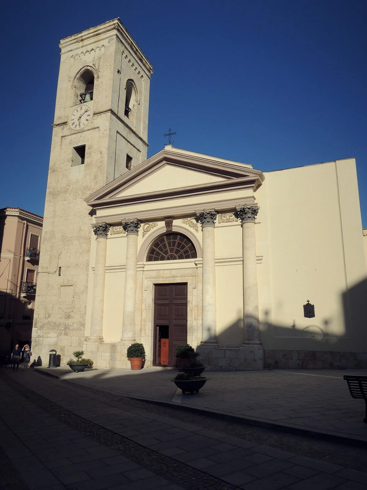 La facciata neoclassica color crema della chiesa di San Giacomo a Cagliari, con colonne corinzie, portone in legno e alto campanile con orologio, sotto un cielo blu intenso
