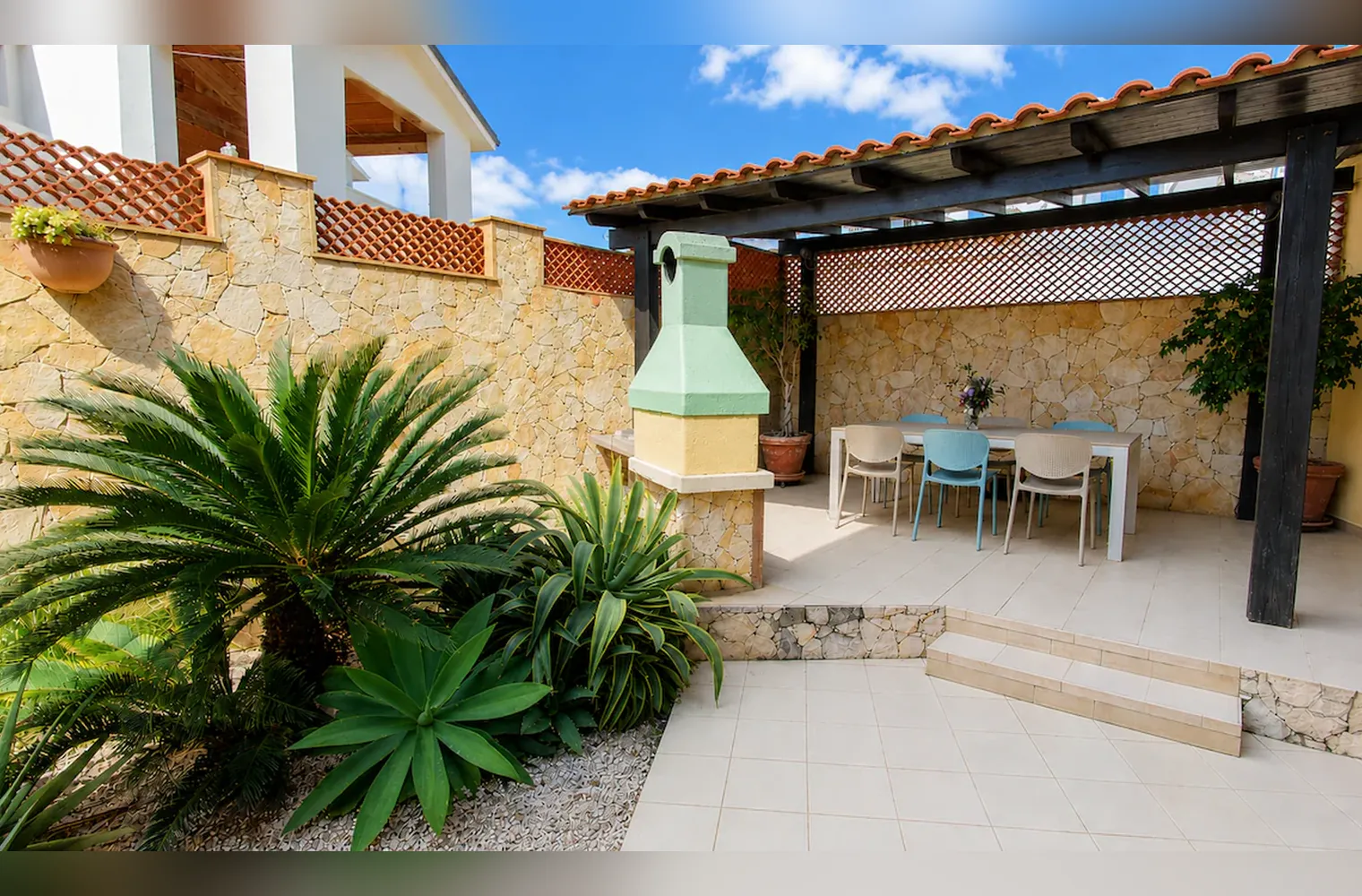 Il giardino con le palme nane e le agavi davanti al pergolato con il tavolo e il barbecue