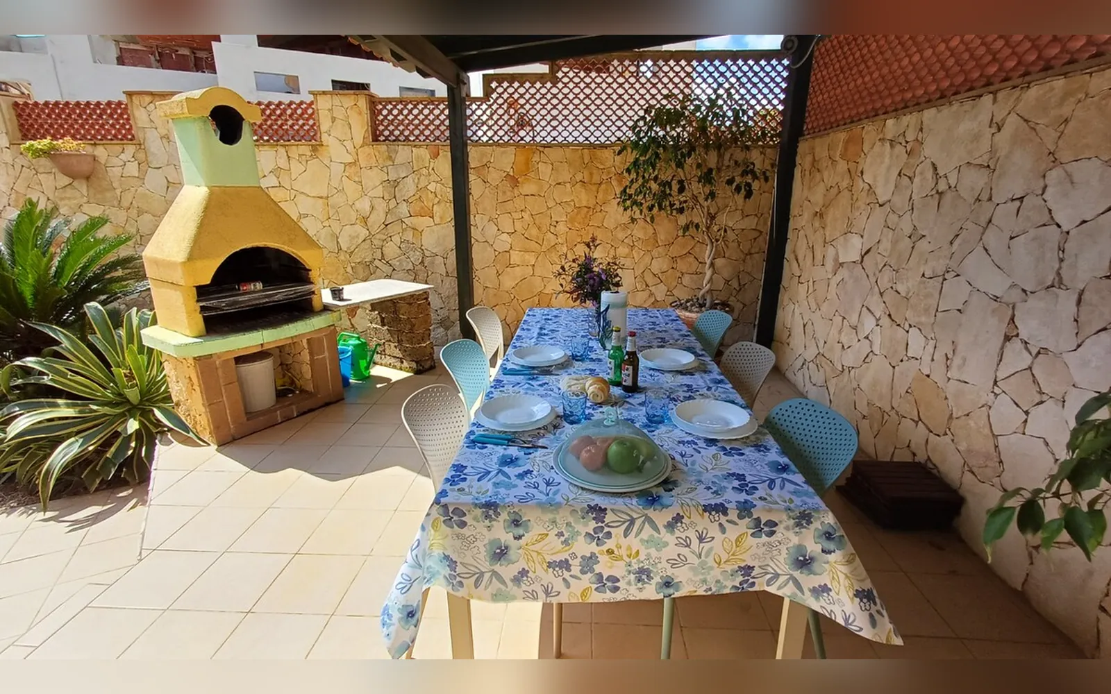 La tavola apparecchiata sotto il pergolato, con il barbecue in muratura e il muro in pietra